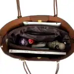 Geschlossene braune Ledertasche mit Innentaschen, gefüllt mit Smartphone, Wasserflasche, Parfüm, Smartphone, Spiele, und Buch - ideal für Reisen und Alltag.