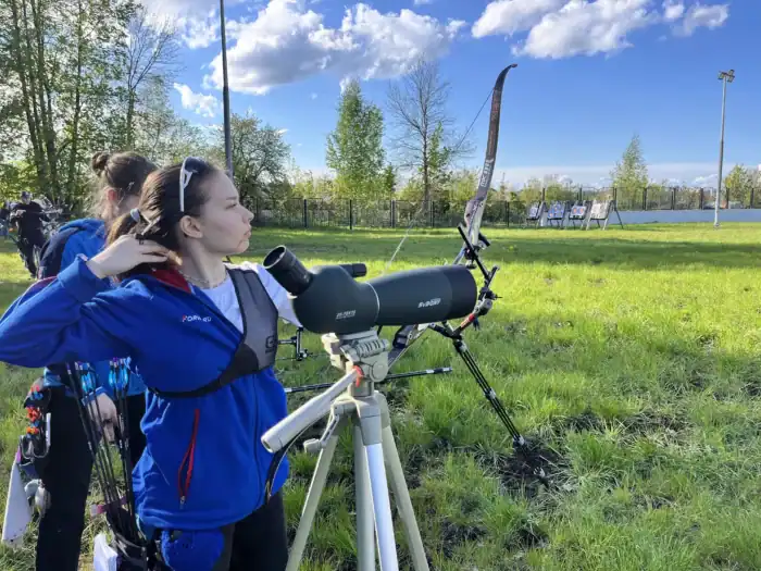 Fernglas auf einem Stativ, junge Bogenschützin, Outdoor Sportschießen, Sportaktivitäten im Grünen, präzises Zieltraining bei schönem Wetter.