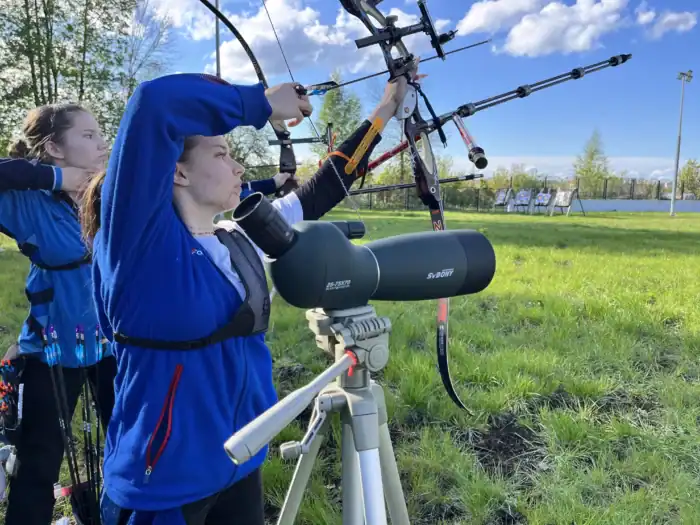 Präzises Sport-Schießen im Freien mit Bogen und Absehen, jungen Athleten beim Training, Outdoor-Sportanlage, optimierte Performance, Fokus auf Genauigkeit und Technik, Sporttruhe für Ausrüstung.